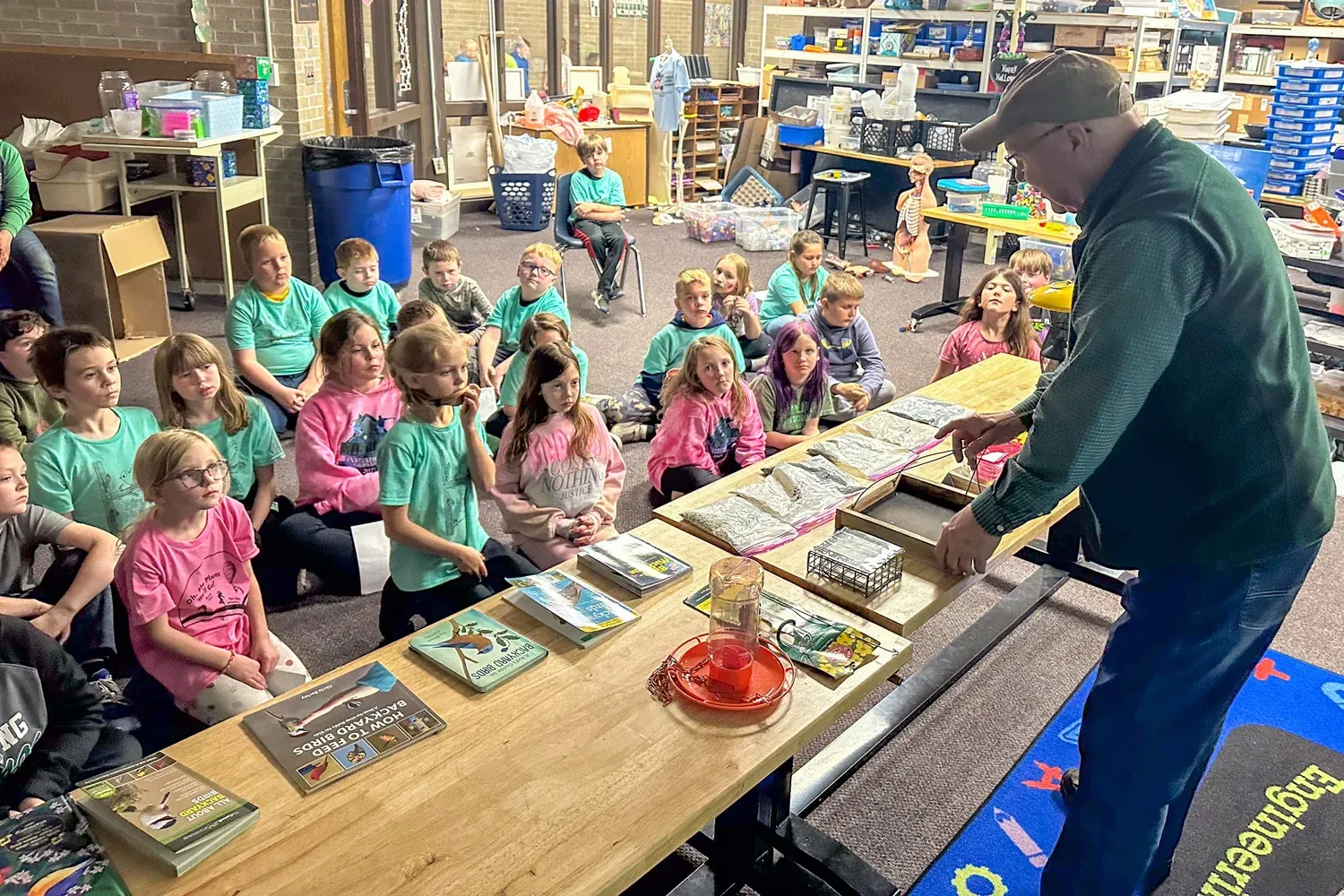 Greg presenting to a large group of seated elementary students, with bird books, a feeder, and nesting materials laid out on a table.