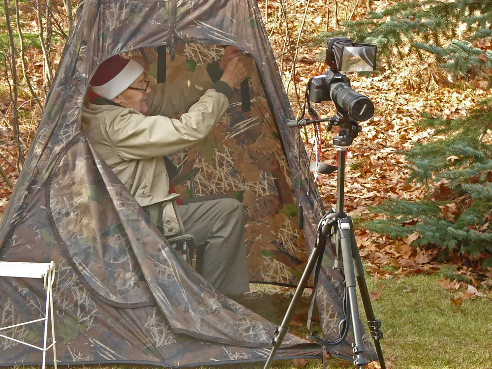 An older man seated with a camera, photographing birds near backyard feeders.