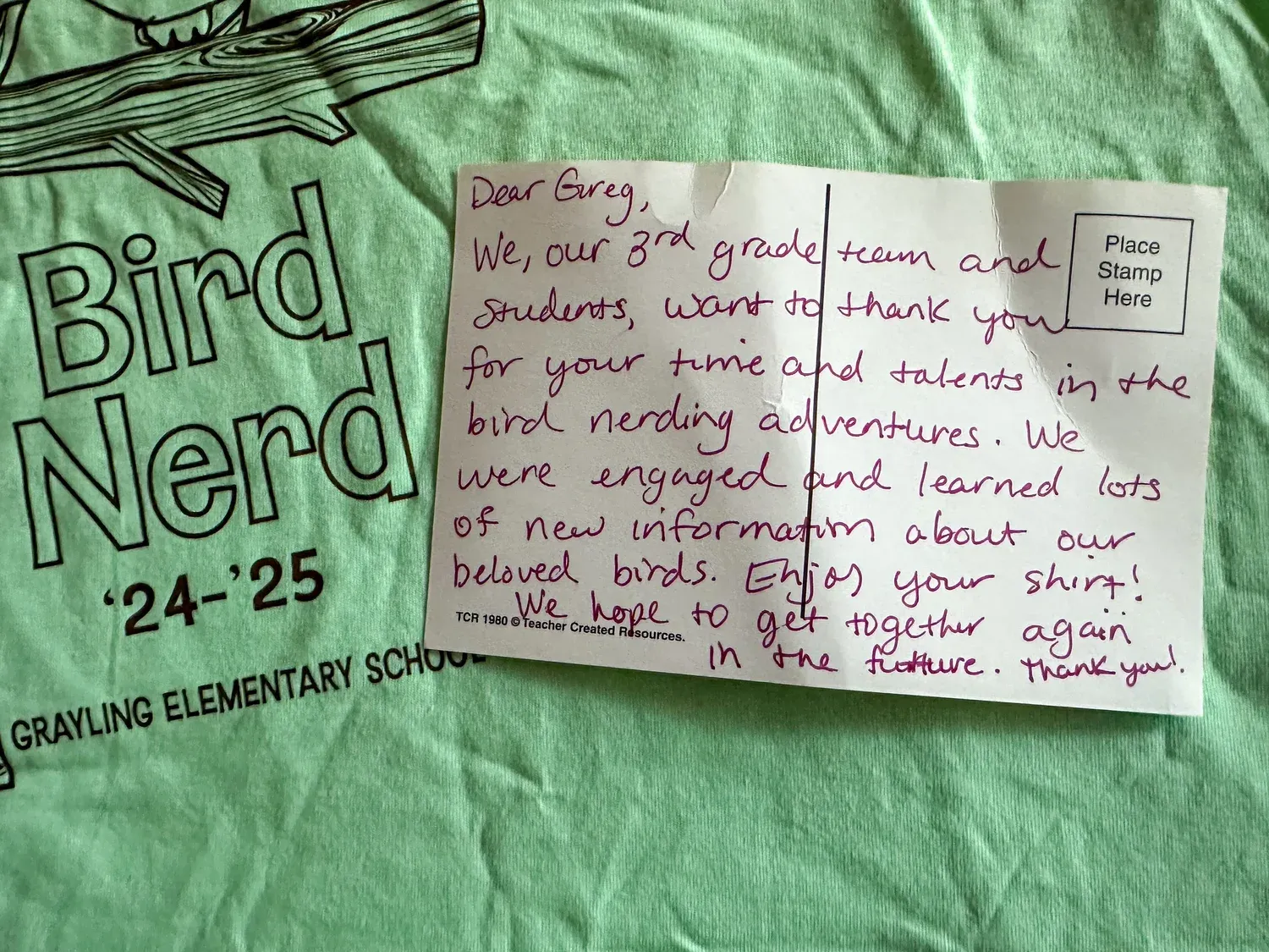 A handwritten thank-you postcard resting on a green “Bird Nerd ’24–’25, Grayling Elementary School” T-shirt. It reads: “Dear Greg, We, our 3rd grade team and students, want to thank you for your time and talents in the bird nerding adventures. We were engaged and learned lots of new information about our beloved birds. Enjoy your shirt! We hope to get together again in the future. Thank you!”
